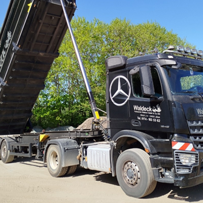 Ein schwarzer Muldenkipper mit dem Waldeck-Logo und dem Mercedes-Benz Emblem entlädt Sand oder Kies auf dem Boden, wobei der Anhänger schräg angehoben ist. Im Hintergrund sind Bäume und ein blauer Himmel zu sehen.