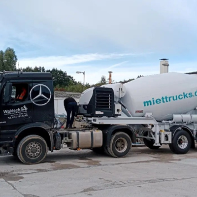 Ein schwarzer Mercedes-Benz Zementmischer mit dem Logo "miettrucks.de" auf der Mischertrommel ist auf einer Betonfläche geparkt. Eine Person arbeitet in der Nähe der Fahrerkabine des LKWs, und im Hintergrund sind Bäume zu sehen.
