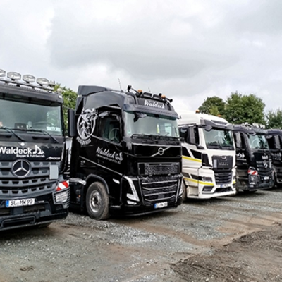 Eine Reihe schwarzer und weißer Sattelschlepper, die auf einem Kiesplatz geparkt sind, mit dem "Waldeck"-Firmenlogo auf den Fahrerhäusern, mit Bäumen und bedecktem Himmel im Hintergrund.