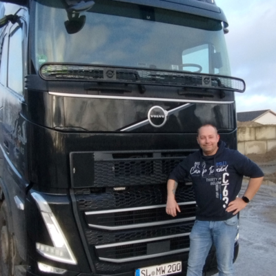 Ein Mann in Freizeitkleidung steht lächelnd mit einem Arm auf der Vorderseite eines großen schwarzen Volvo-Lkw mit der Aufschrift Waldeck. Der Lkw ist auf einer Park-Fläche mit Gebäuden im Hintergrund geparkt.