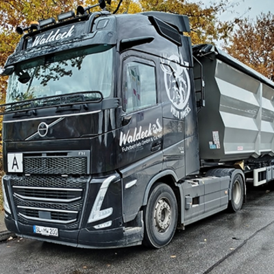 Ein schwarzer Volvo-Sattelzug mit dem Namen Waldeck auf dem Führerhaus parkt auf einer nassen Straße neben Herbstbäumen und zieht einen großen grauen Anhänger.