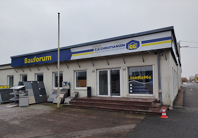 Ein Gebäude mit den Schildern Bauforum und C.G. Christiansen steht auf einem gepflasterten Grundstück. Draußen stehen Schautafeln und Baumaterialien sowie Schilder mit Renovierungsangeboten. In der Nähe des Eingangs befindet sich ein orangefarbener Verkehrskegel.