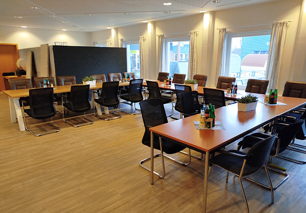 Ein moderner Konferenzraum mit mehreren langen, U-förmig angeordneten Tischen, umgeben von schwarzen Stühlen. Auf den Tischen stehen Getränkeflaschen, Gläser und Topfpflanzen. Große Fenster lassen natürliches Licht herein.