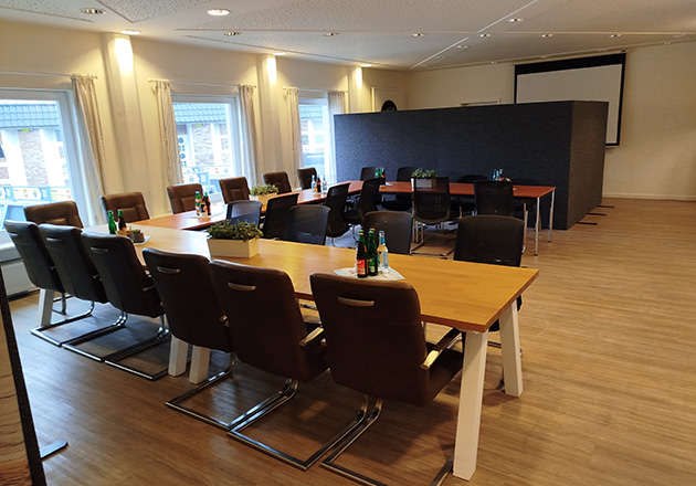 Ein Konferenzraum mit zwei langen Holztischen, die in U-Form angeordnet sind, umgeben von braunen Stühlen. Auf den Tischen stehen Wasserflaschen, Gläser und kleine Topfpflanzen. Große Fenster lassen natürliches Licht herein.